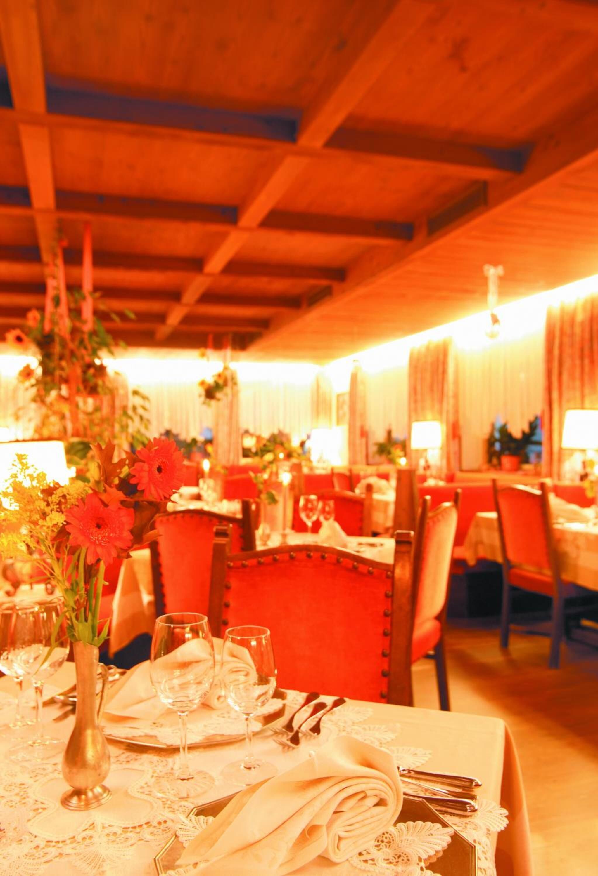 A warmly lit dining area with tables set for a meal, featuring floral centerpieces and red upholstered chairs.