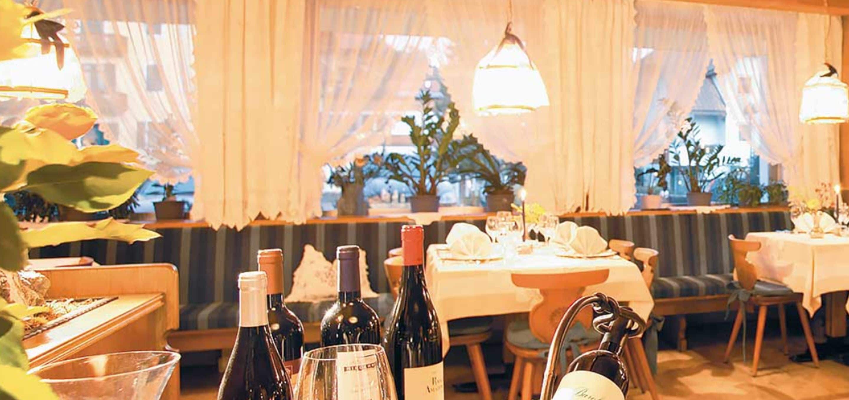 Un restaurant où les tables sont dressées pour le repas, avec des plantes en pot et des bouteilles de vin posées dessus.