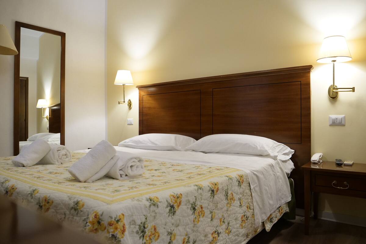 A hotel room with a wooden headboard, a bed with white linens, two wall-mounted lamps, a bedside table with a phone, and a mirror.