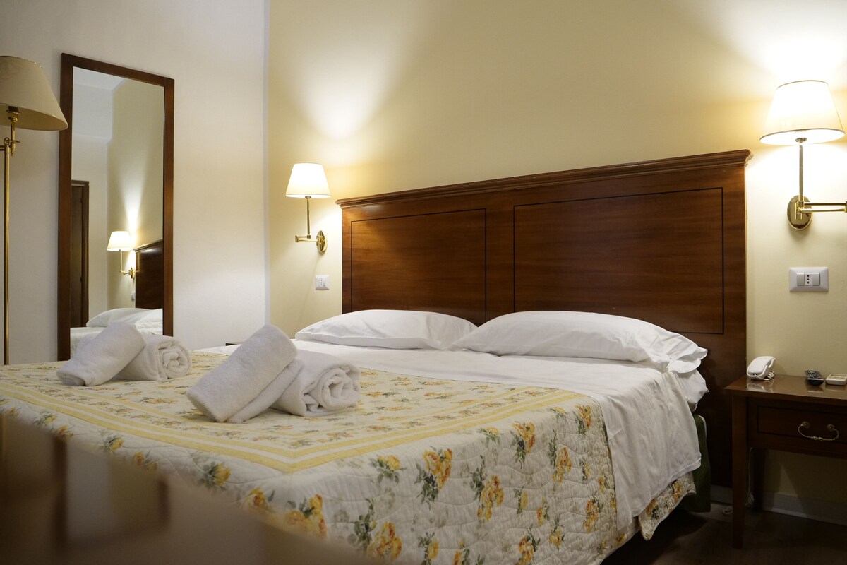A hotel room with a large bed, two bedside lamps, a wooden headboard, a nightstand with a phone, and a mirror.