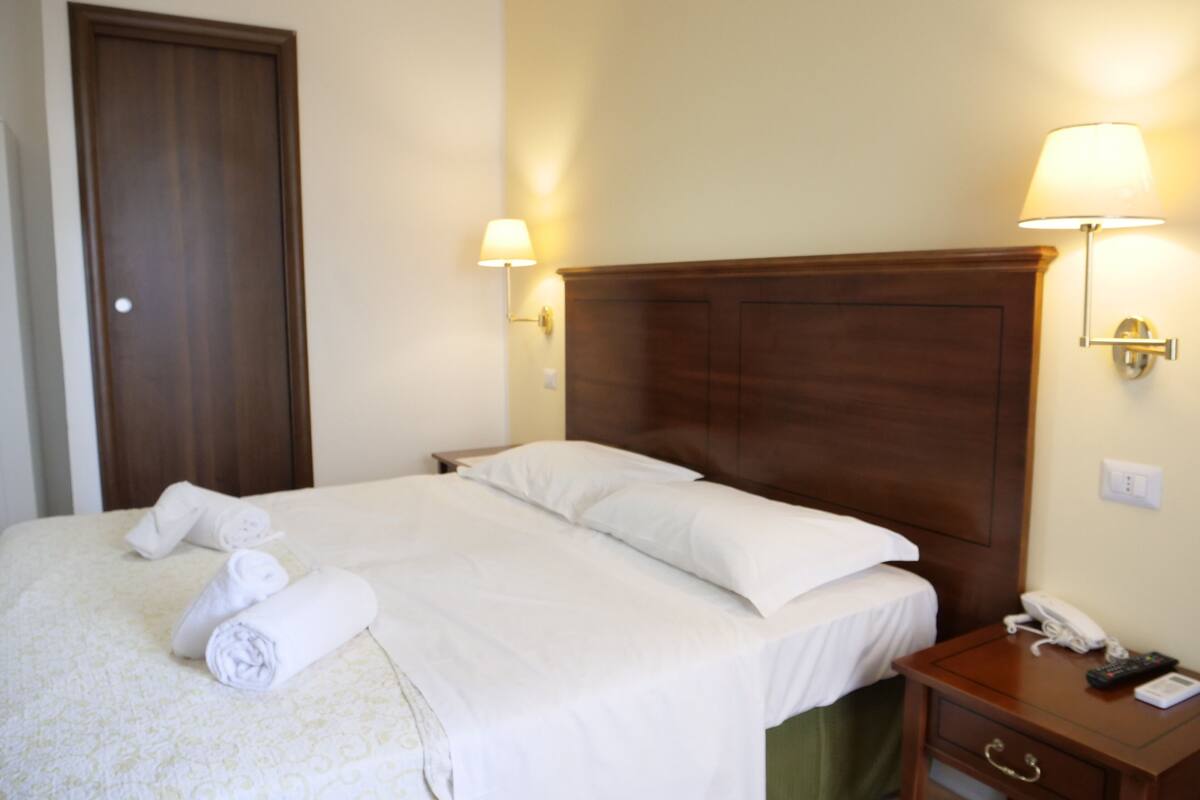 A hotel room with a large bed, two bedside lamps, a wooden headboard, a bedside table with a phone, and a door.