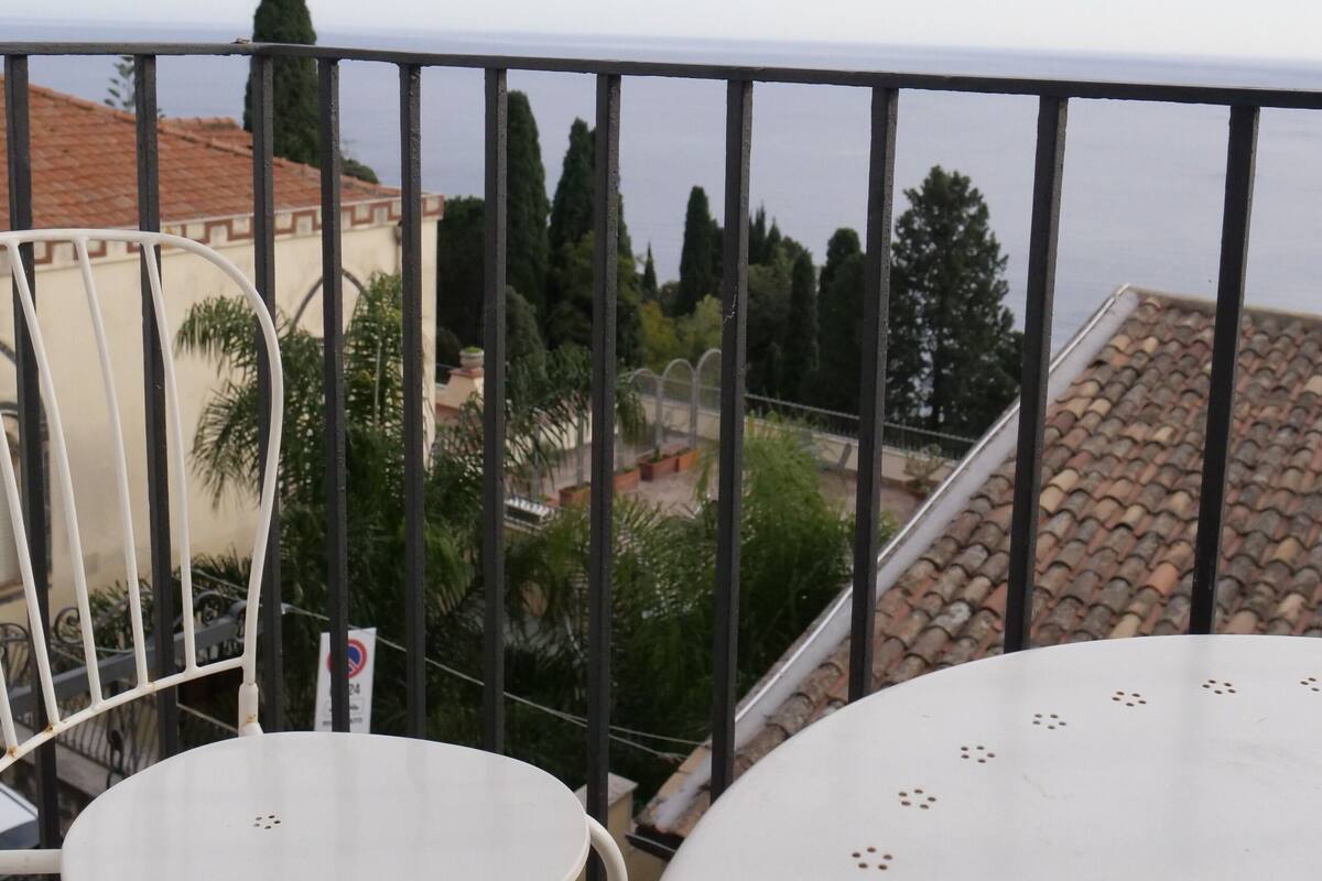 A balcony with a white table and chair, overlooking a coastal area with buildings and trees.