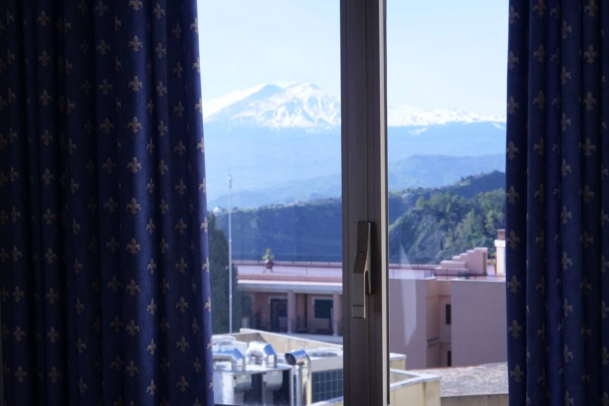 A view through a window with curtains, featuring a mountain range and buildings in the background.