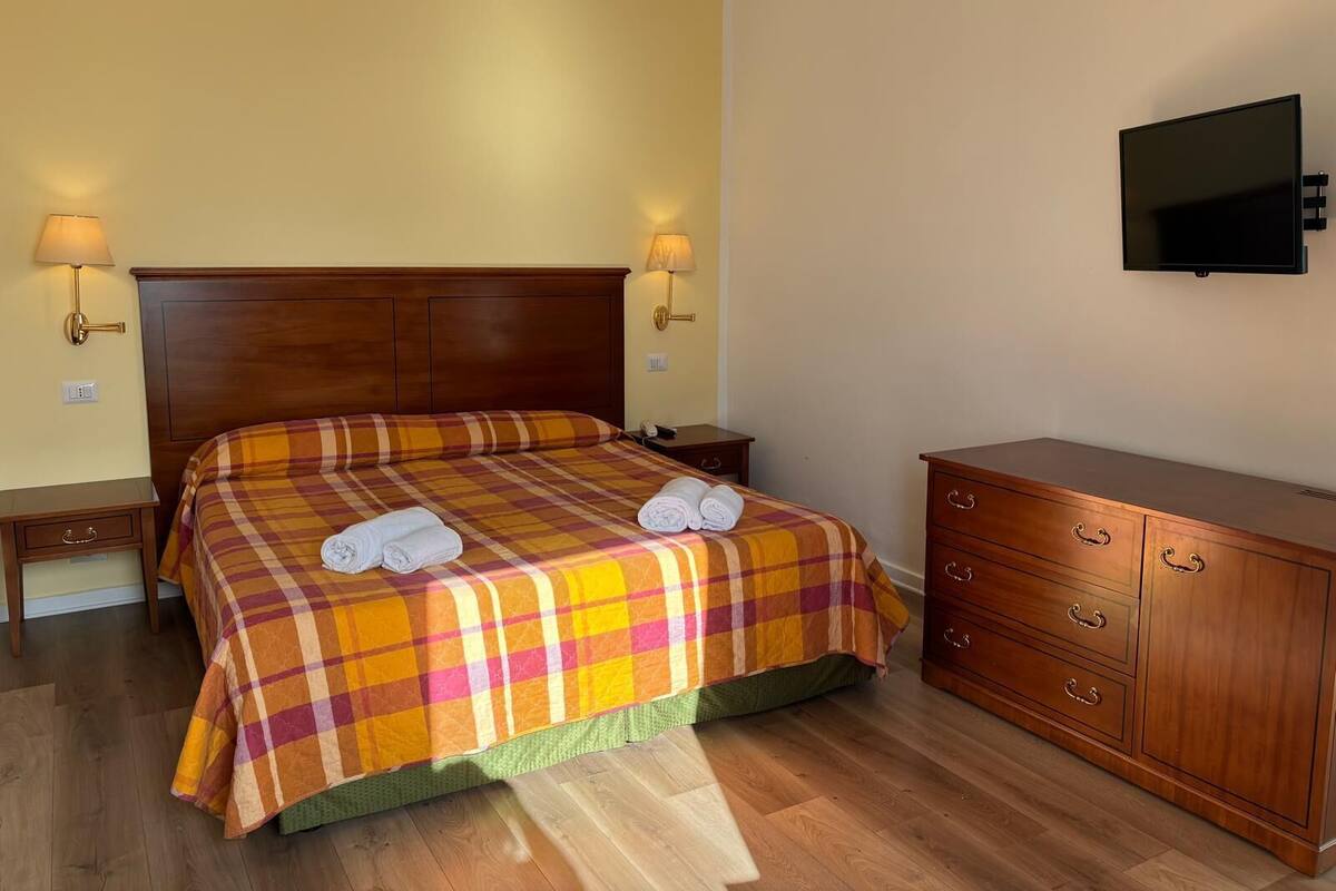 A hotel room with a wooden bed, two bedside tables, a TV mounted on the wall, and a wooden dresser.