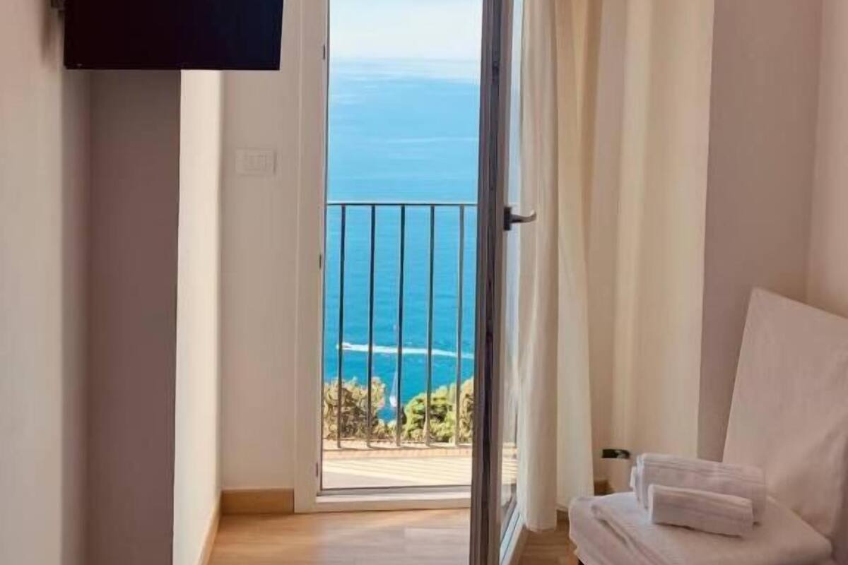 A narrow hallway leading to a balcony with a sea view, a mounted flat-screen TV, and a chair with towels.