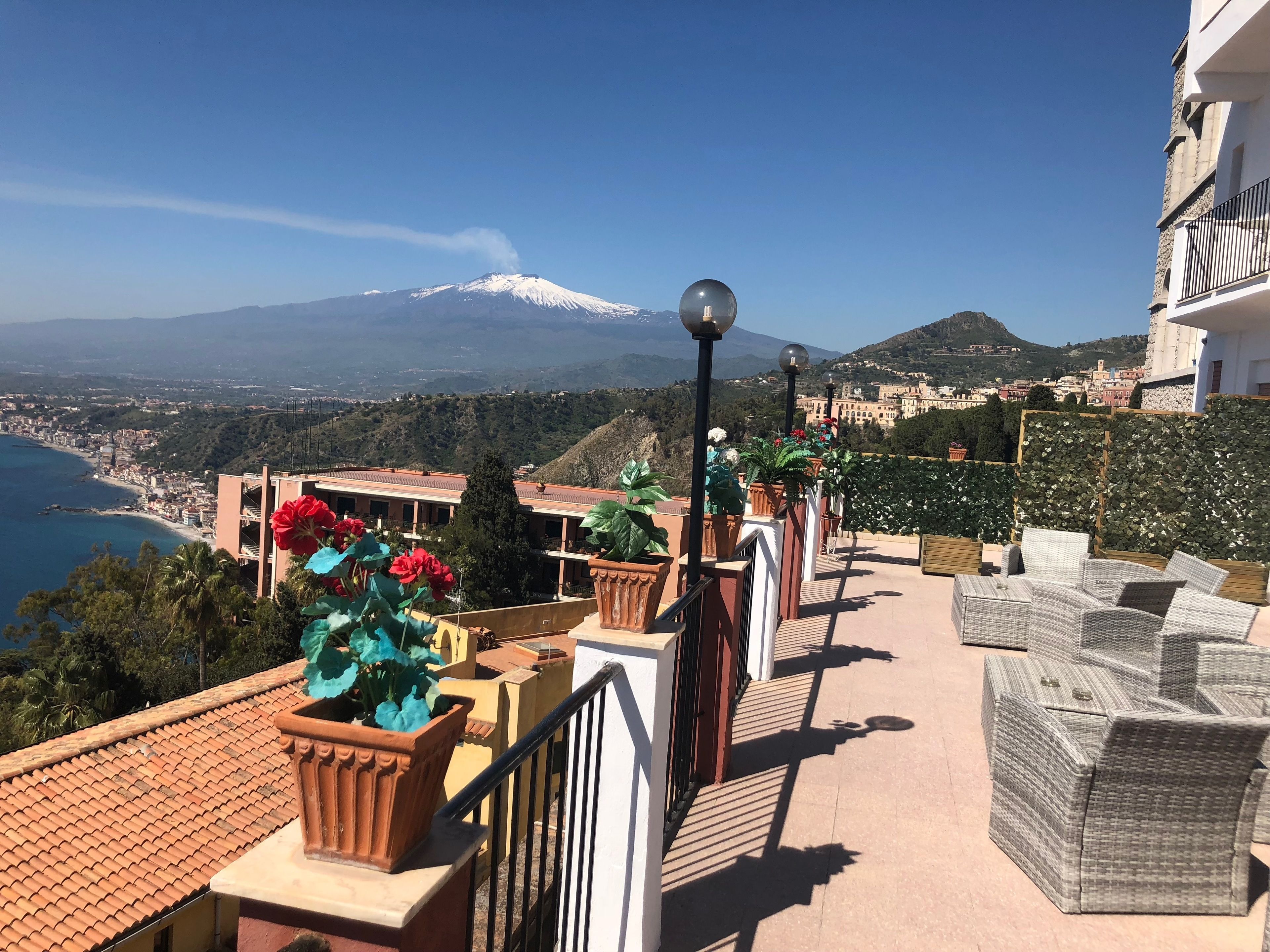 A balcony with wicker furniture, potted plants, and a view of a snow-capped mountain and a coastal town.