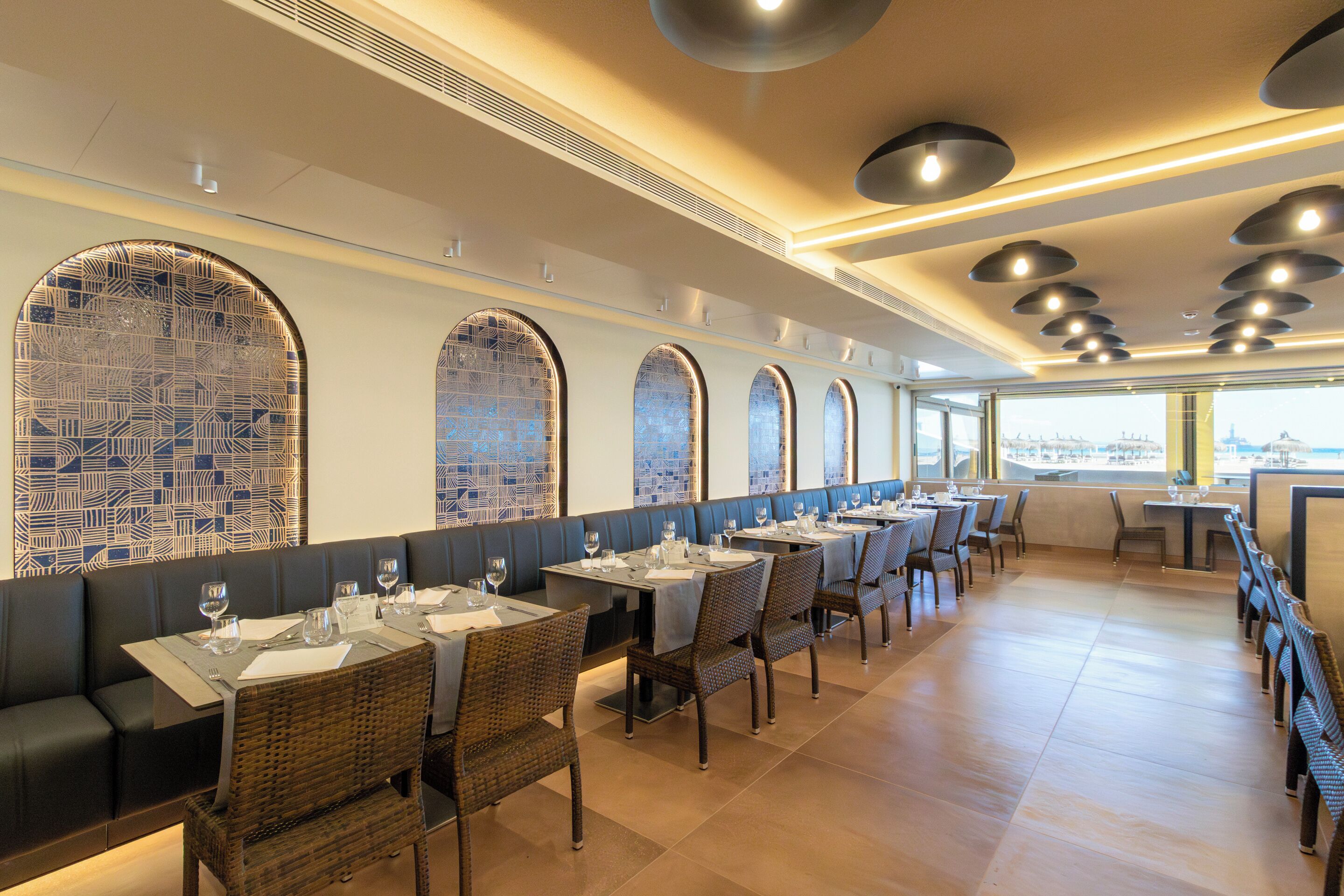 Un restaurante con mesas largas, sillas de mimbre y ventanas en arco.