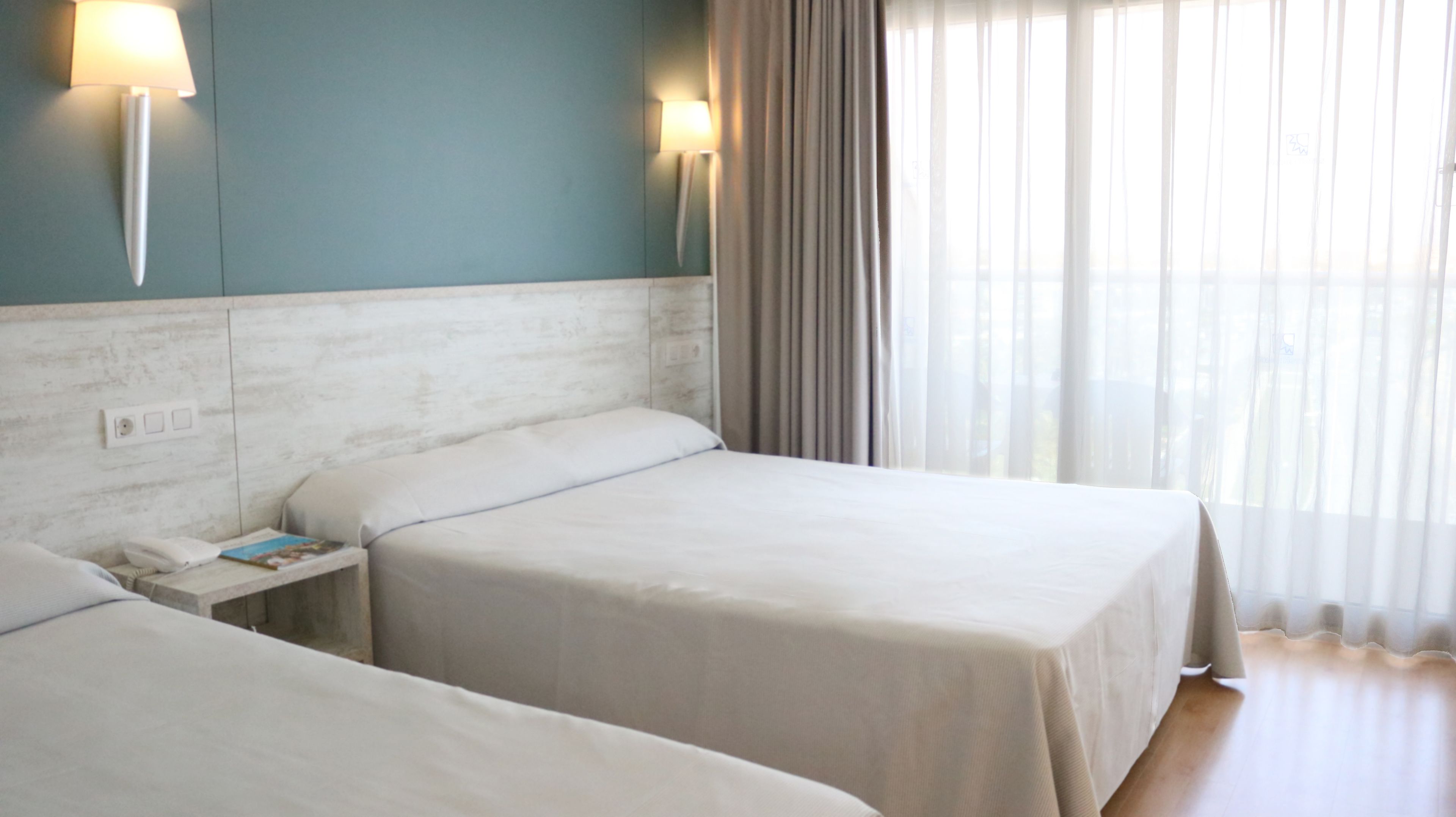A hotel room with two beds, a nightstand, a lamp, and a window with curtains.