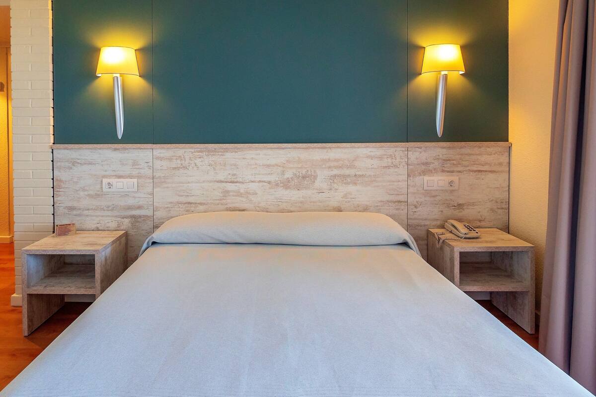 Un lit bien fait, recouvert d’une couvre-lit à motifs, avec deux lampes de chevet, une tête de lit en bois et une petite table de chevet sur laquelle se trouve un téléphone.