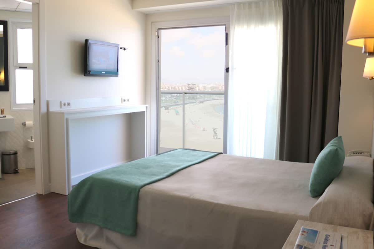 Une chambre d’hôtel avec un lit, un téléviseur fixé au mur, une fenêtre donnant sur la plage, et une salle de bain avec un miroir et un lavabo.