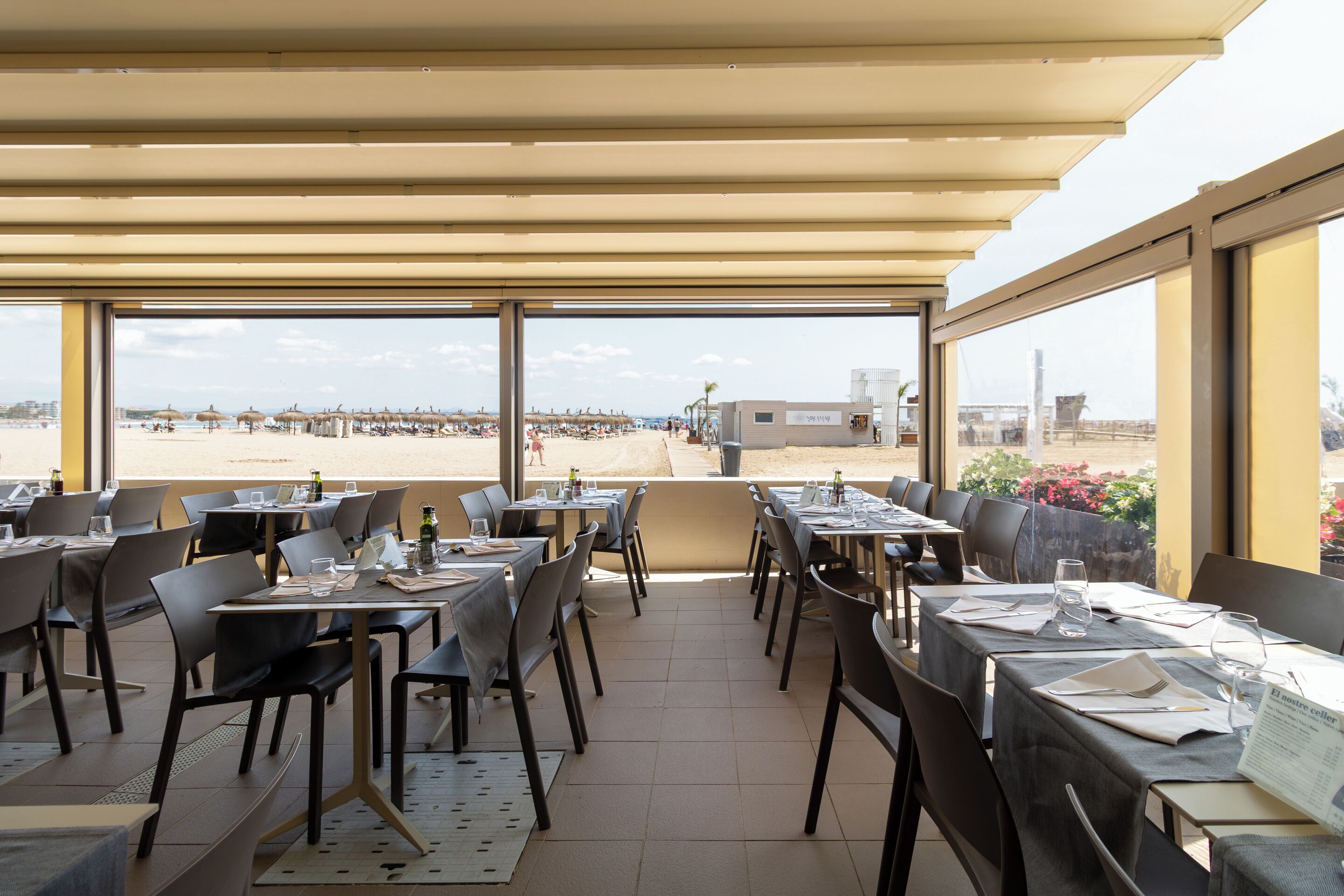 Un área de comedor al aire libre con mesas y sillas, vista a la playa y un edificio al fondo.