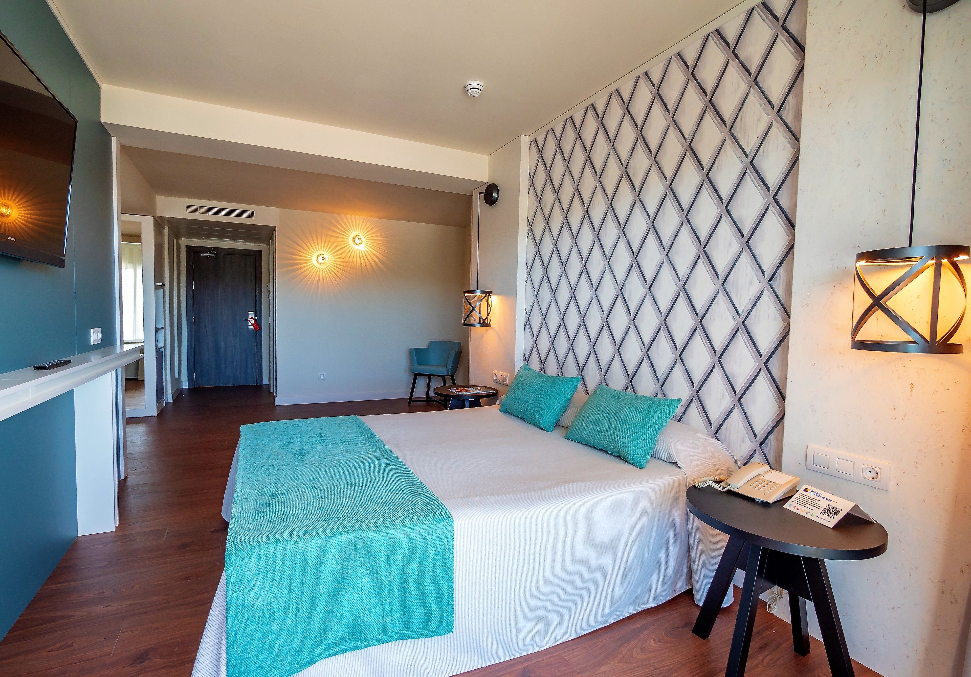 A hotel room with a large bed, a desk with a telephone, and a wall-mounted light fixture.