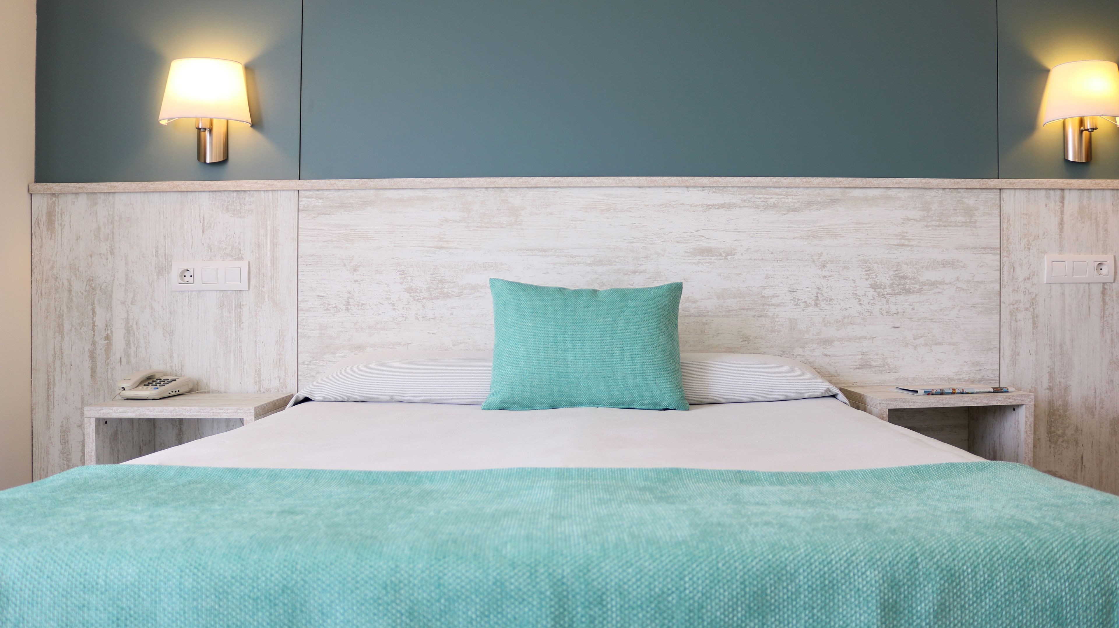 A hotel room with a bed, a teal pillow, two bedside lamps, and a telephone on a nightstand.
