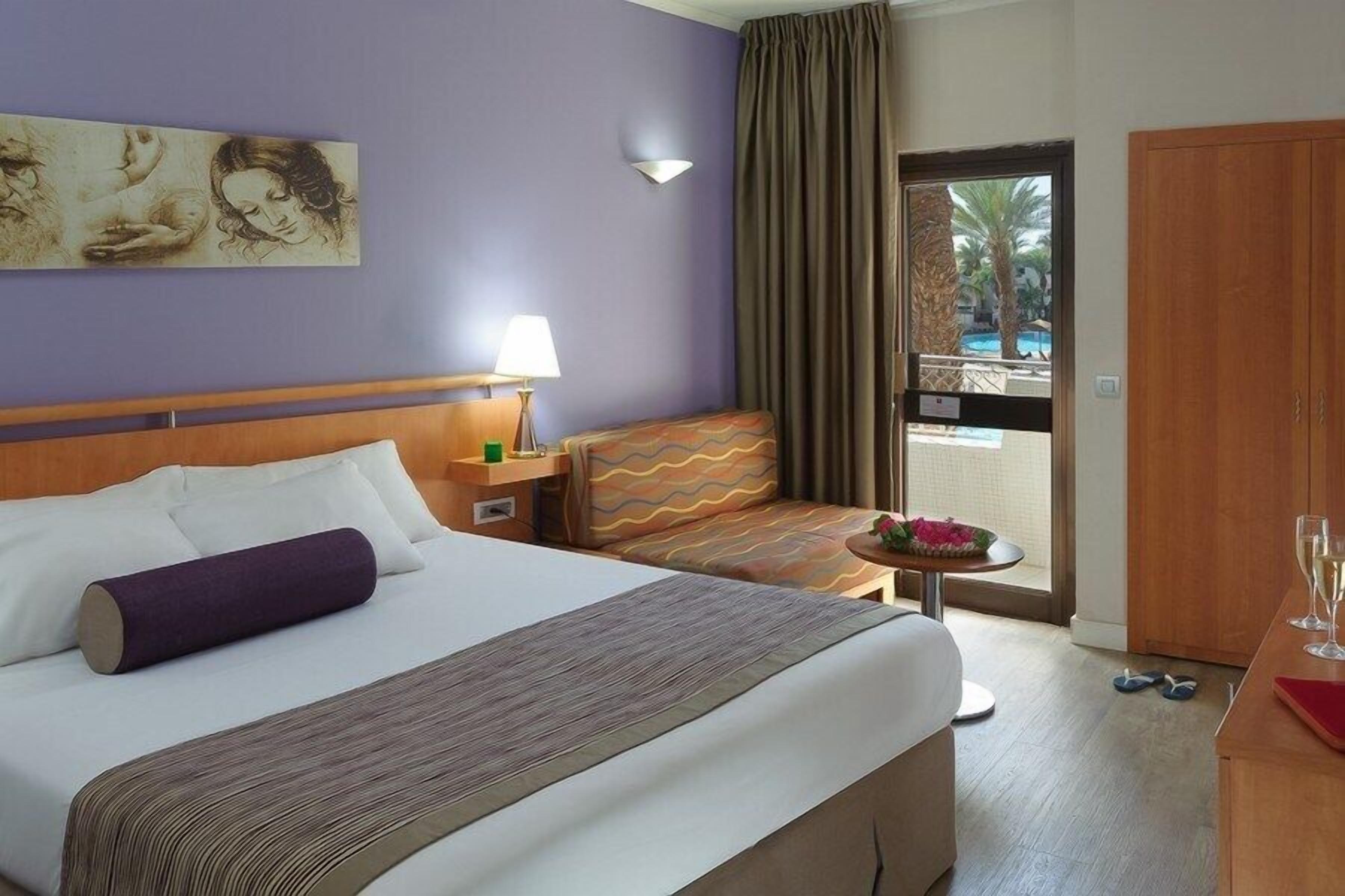 A hotel room with a bed, a sofa, a small table with a lamp, a window with curtains, and a door leading to a balcony.