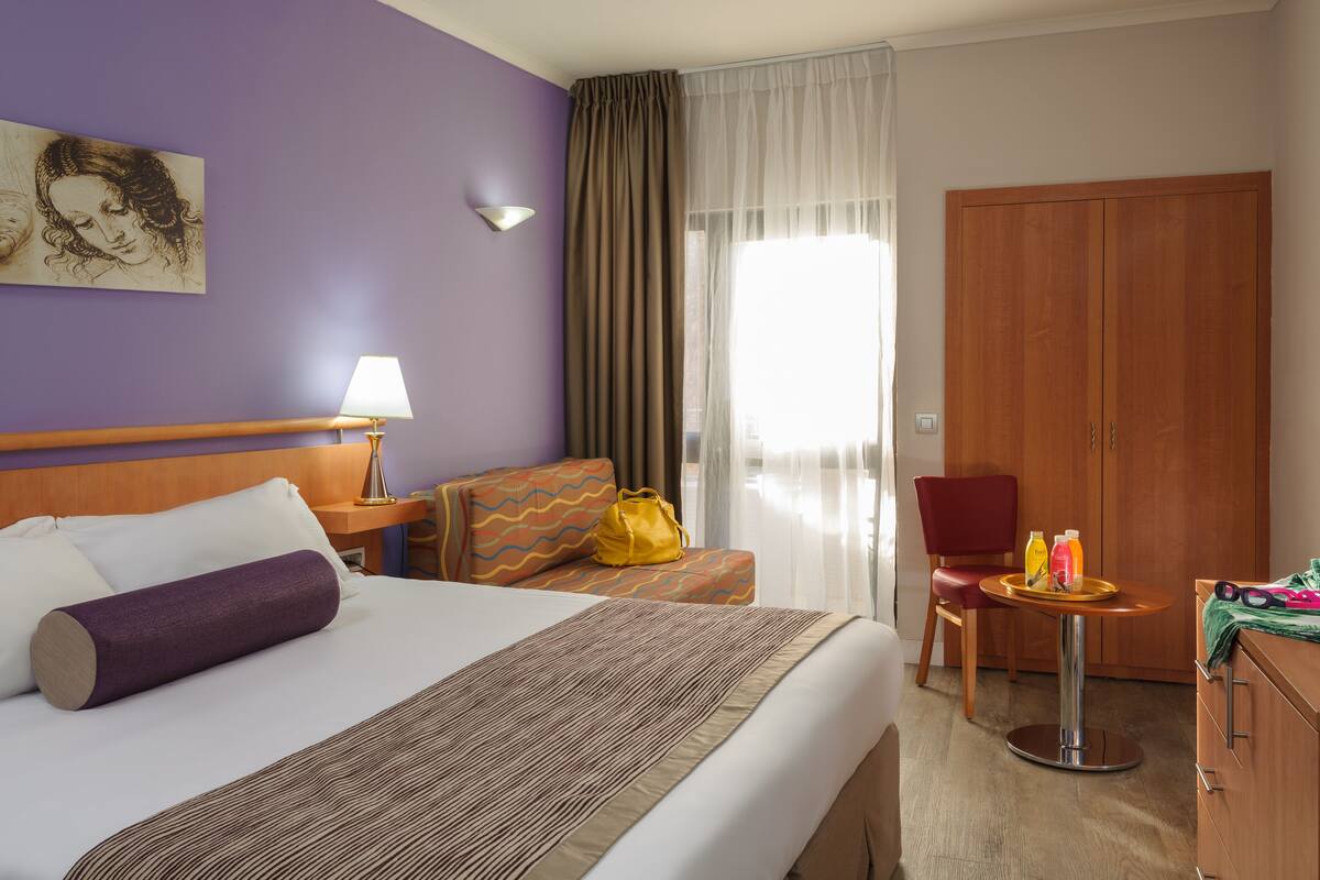 A hotel room with a bed, a desk, a chair, a wardrobe, and a window with curtains.