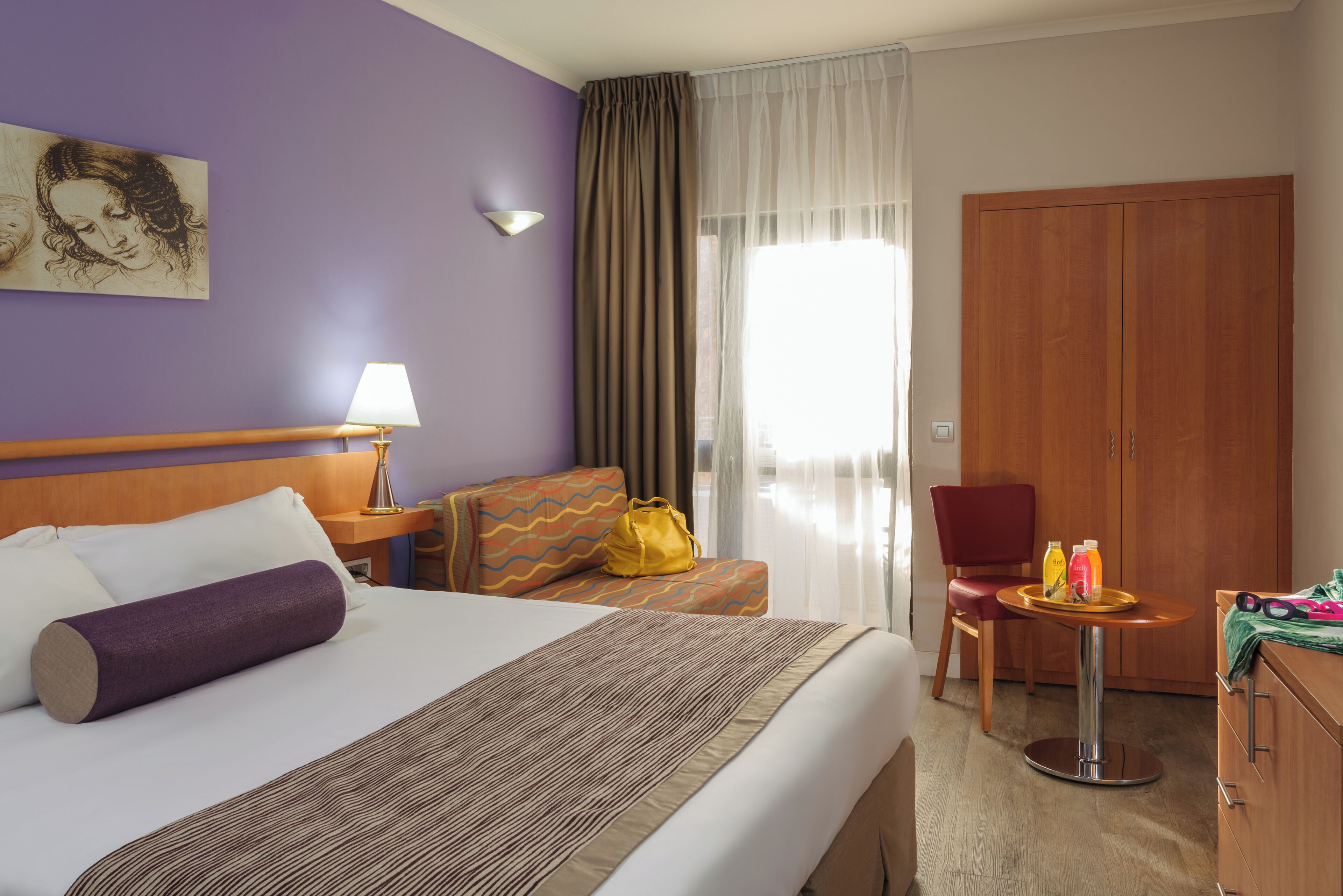 A hotel room with a bed, a desk, a chair, a wardrobe, and a window with curtains.