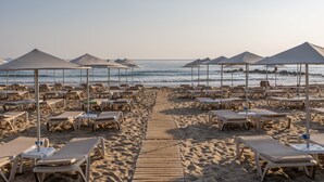 Une plage bordée de rangées de chaises longues et de parasols, une promenade en bois menant à l’eau, et une vue dégagée sur l’océan.