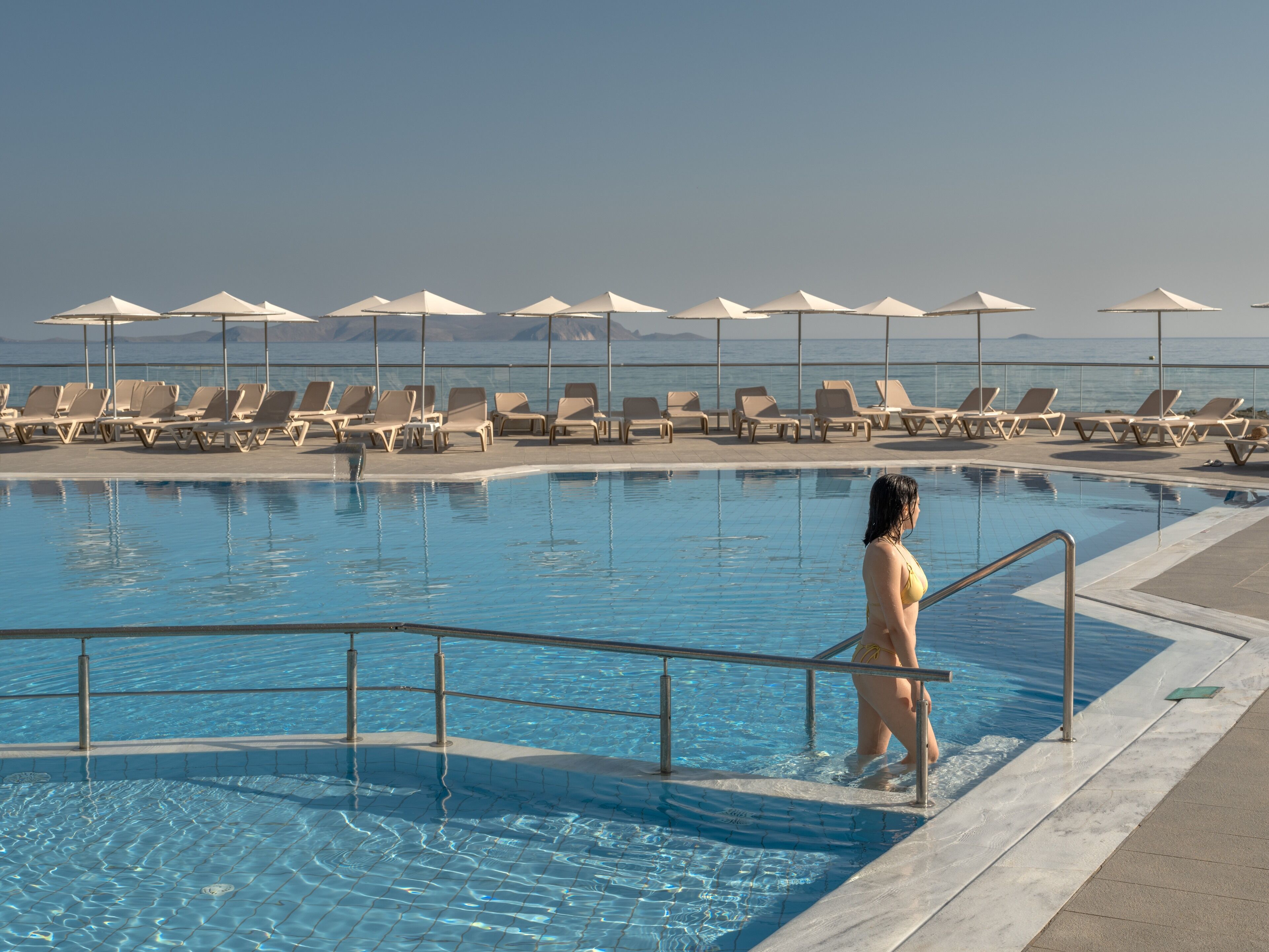Zona de piscina con sillones de descanso y sombrillas, una mujer en traje de baño y una vista despejada del mar al fondo.
