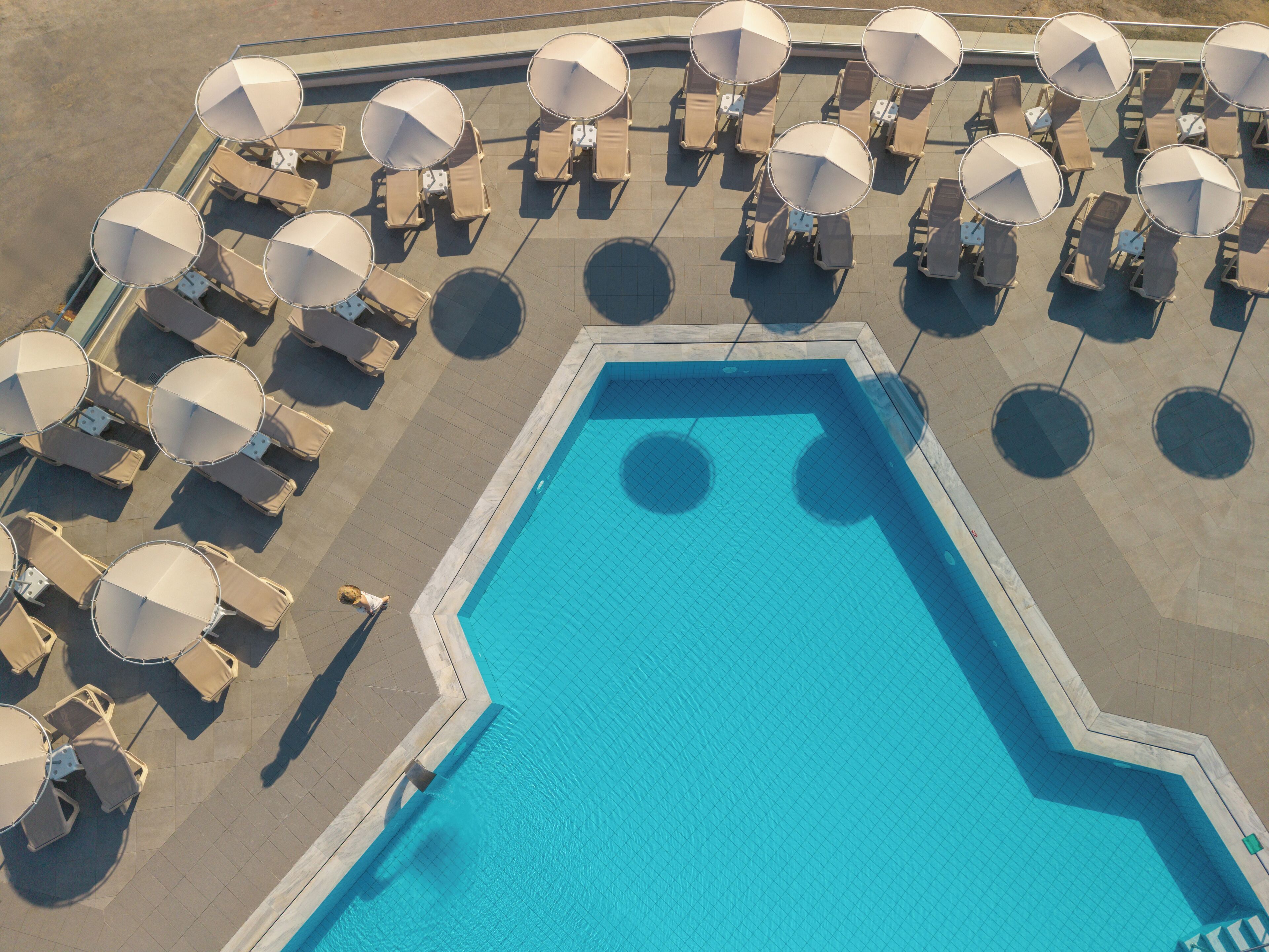 An aerial view of a swimming pool surrounded by lounge chairs and umbrellas, with a tiled deck and a clear blue pool.