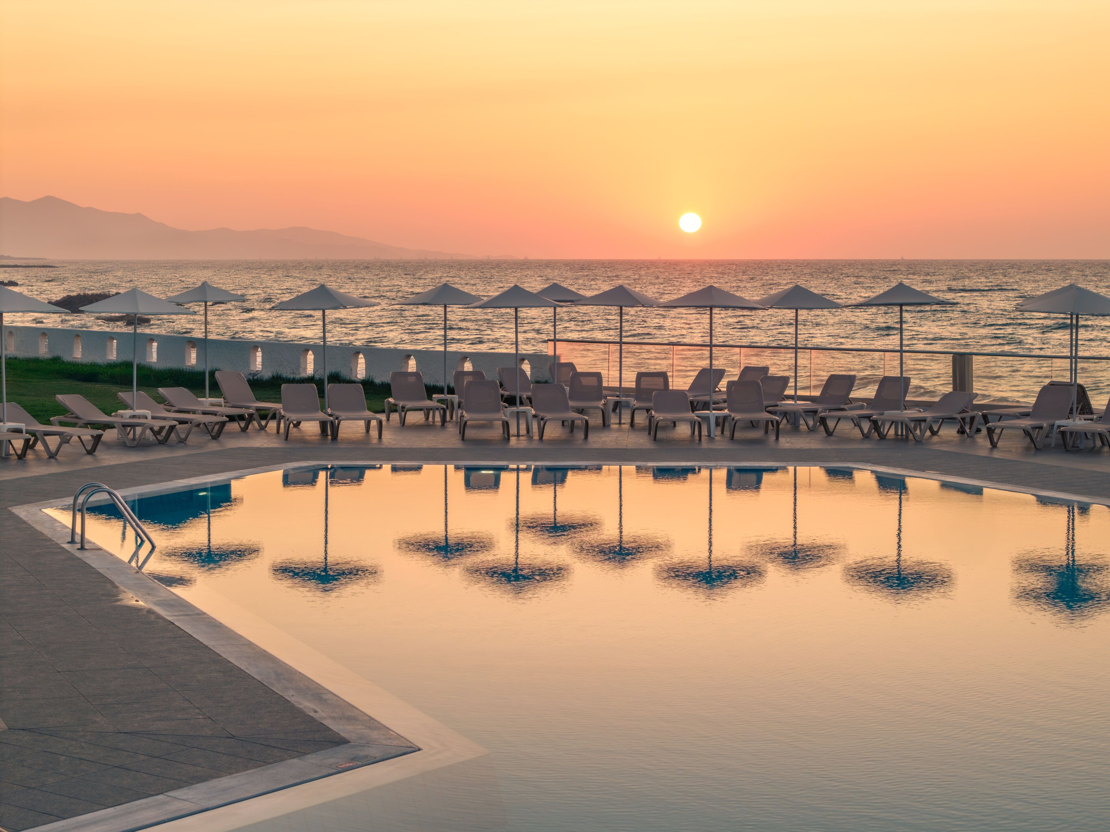 Un espace aménagé au bord de la piscine, avec des chaises longues et des parasols, offrant une vue sur une étendue d’eau au coucher du soleil.