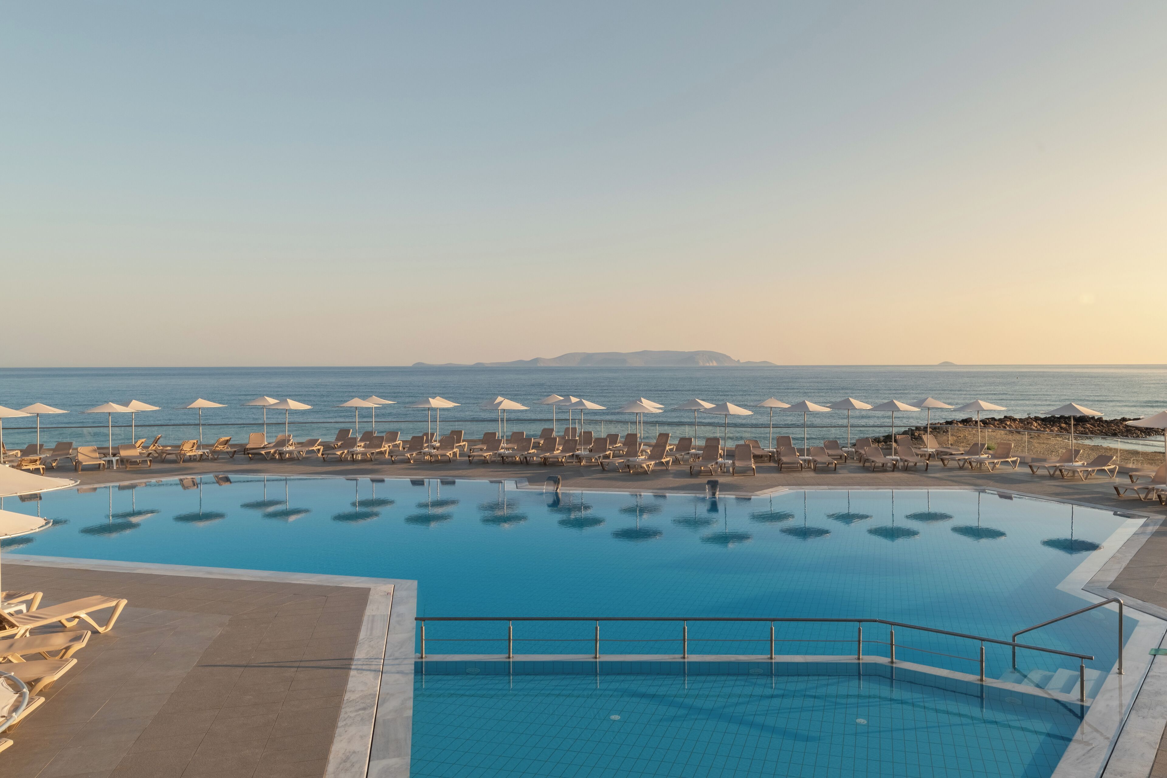 Une grande piscine bordée de chaises longues et d’ombrelles, offrant une vue dégagée sur la mer et les îles lointaines.