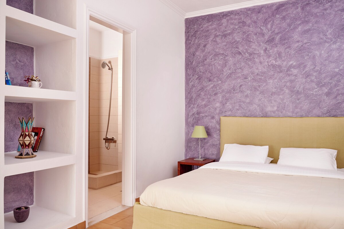 A bedroom with a bed, a nightstand, a shelf with decorative items, and a shower area.