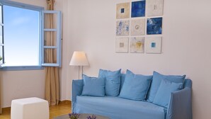 A modern living room with a blue sofa, two white poufs, a round coffee table with potted plants, a window with curtains, and a wall with abstract art. - Manos Small World (Santorini)