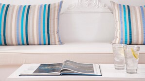 A white sofa with striped pillows, a magazine, and two glasses of water with lemon slices on a white table. - Manos Small World (Santorini)