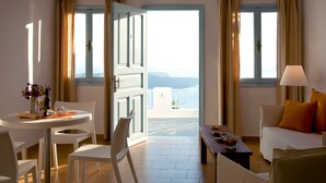 A room with a dining table, chairs, a sofa, and a view of the sea through open doors. - Manos Small World (Santorini)