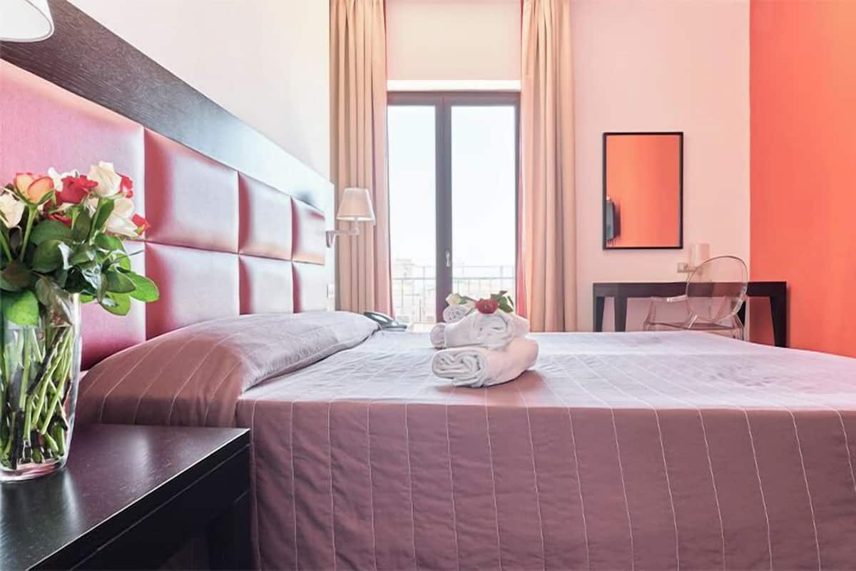 A hotel room with a bed, a nightstand, a vase of flowers, and a window with curtains.