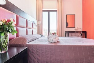 Una habitación de hotel con cama, mesita de noche, un jarrón de flores y ventana con cortinas.