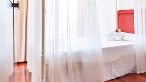 Una habitación de hotel con piso de madera, una cama con cabecera roja y cortinas blancas traslúcidas.