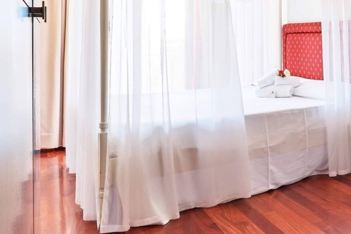 A hotel room with a wooden floor, a bed with a red headboard, and sheer white curtains.