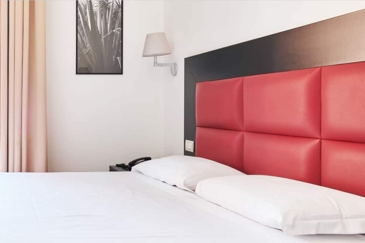 A neatly made bed with white linens, a red upholstered headboard, and a wall-mounted lamp.