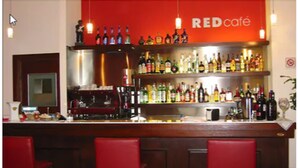 Zona de bar con un letrero rojo de café, estantes llenos de botellas de licores, una máquina de café y taburetes de bar rojos.
