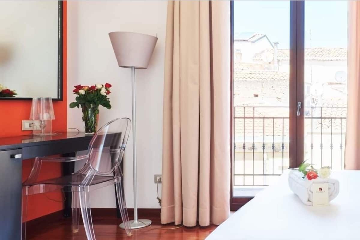 A hotel room with a bed, a desk with a chair, a lamp, and a window with curtains.
