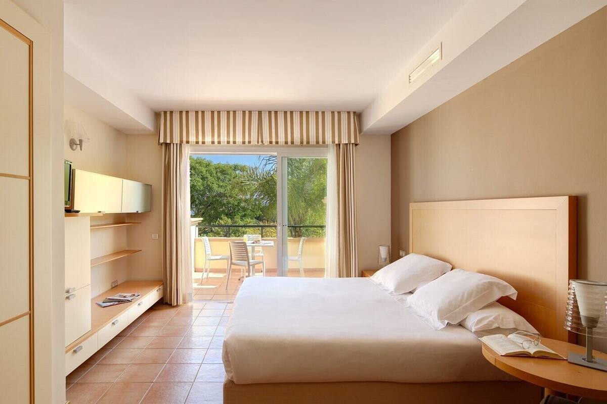 Une chambre d’hôtel avec un grand lit, un téléviseur fixé au mur, un balcon avec des chaises et une armoire en bois.