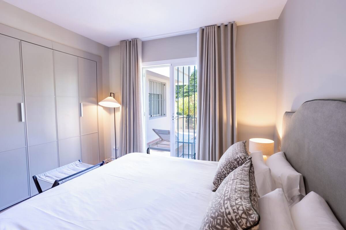 Une chambre d’hôtel avec un grand lit, une armoire, un balcon avec une chaise et une vue sur la verdure environnante.