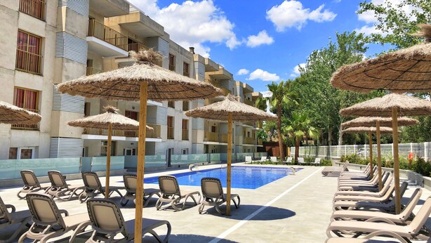 A poolside area with lounge chairs and thatched umbrellas, a clear blue sky, and a building with balconies. - Apartamentos Pins Marina Cambrils (Cambrils)