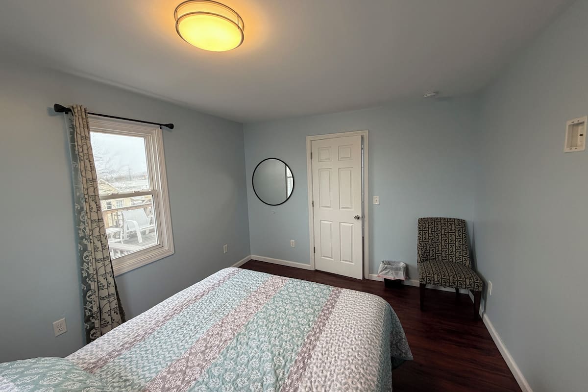 A bedroom with a bed, a window with curtains, a door, a chair, and a round mirror on the wall.