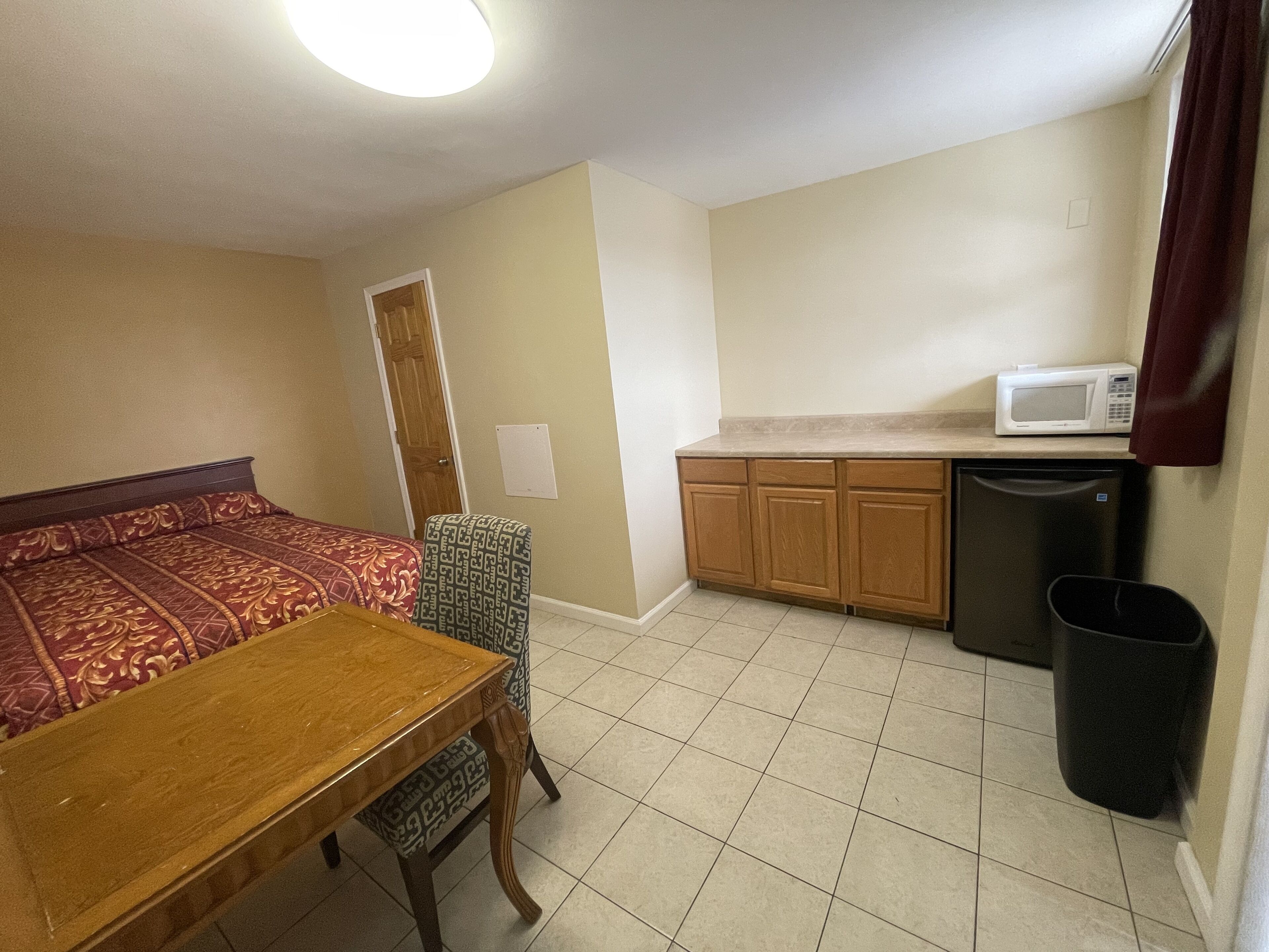 A compact room with a bed, a wooden table, a kitchenette with a microwave, and a trash bin.
