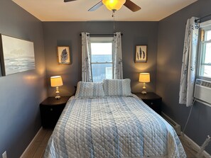 A bedroom with a bed, two bedside tables with lamps, a ceiling fan, a window with curtains, and framed artwork on the walls. - Anchor Motel (Seaside Heights)