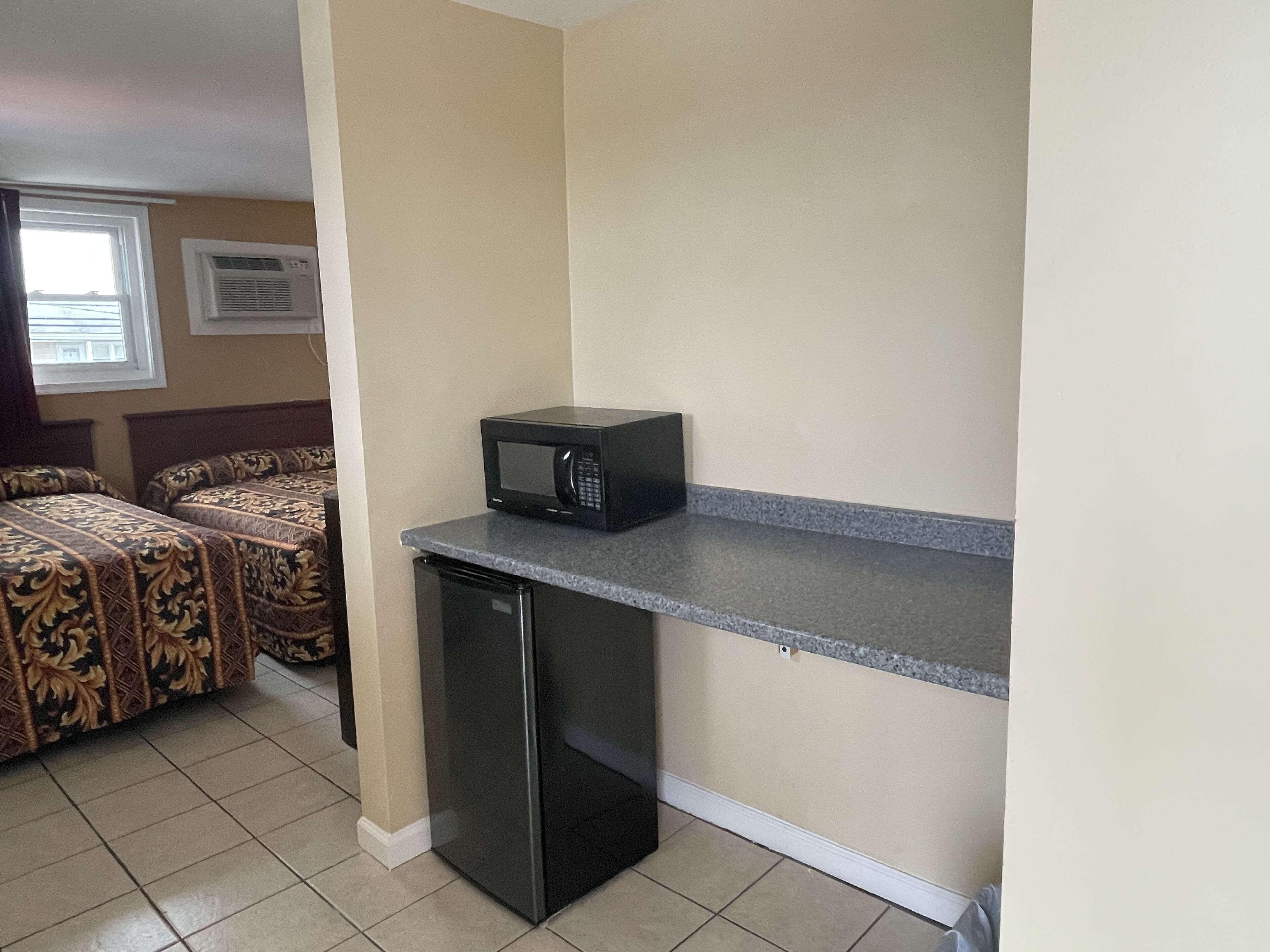 A compact kitchenette with a microwave and a small refrigerator. There are two beds with patterned bedding in the background.