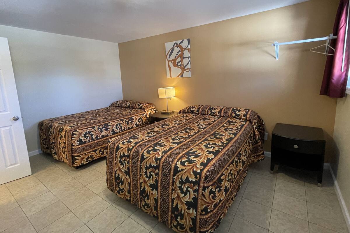 A room with two beds, a nightstand, a lamp, and a hanging coat rack.