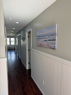 A hallway with wooden flooring, white wainscoting, and a painting of a seascape on the wall.