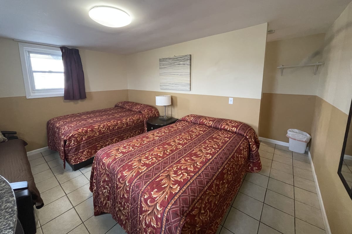 A hotel room with two beds, a small desk, a window with curtains, a ceiling light, and a wall-mounted artwork.