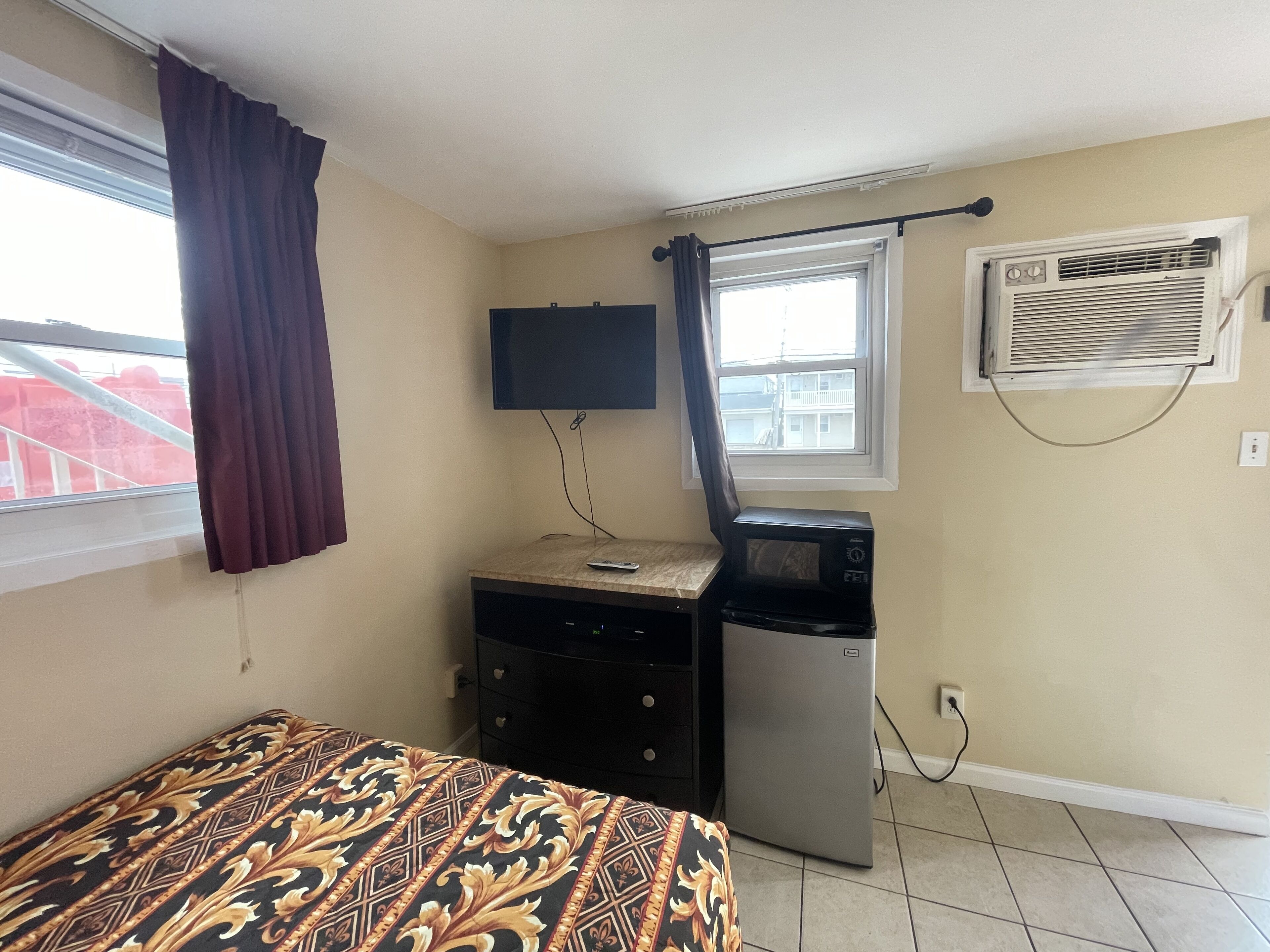A small, sparsely furnished room with a bed, a desk, a microwave, and a window with curtains.