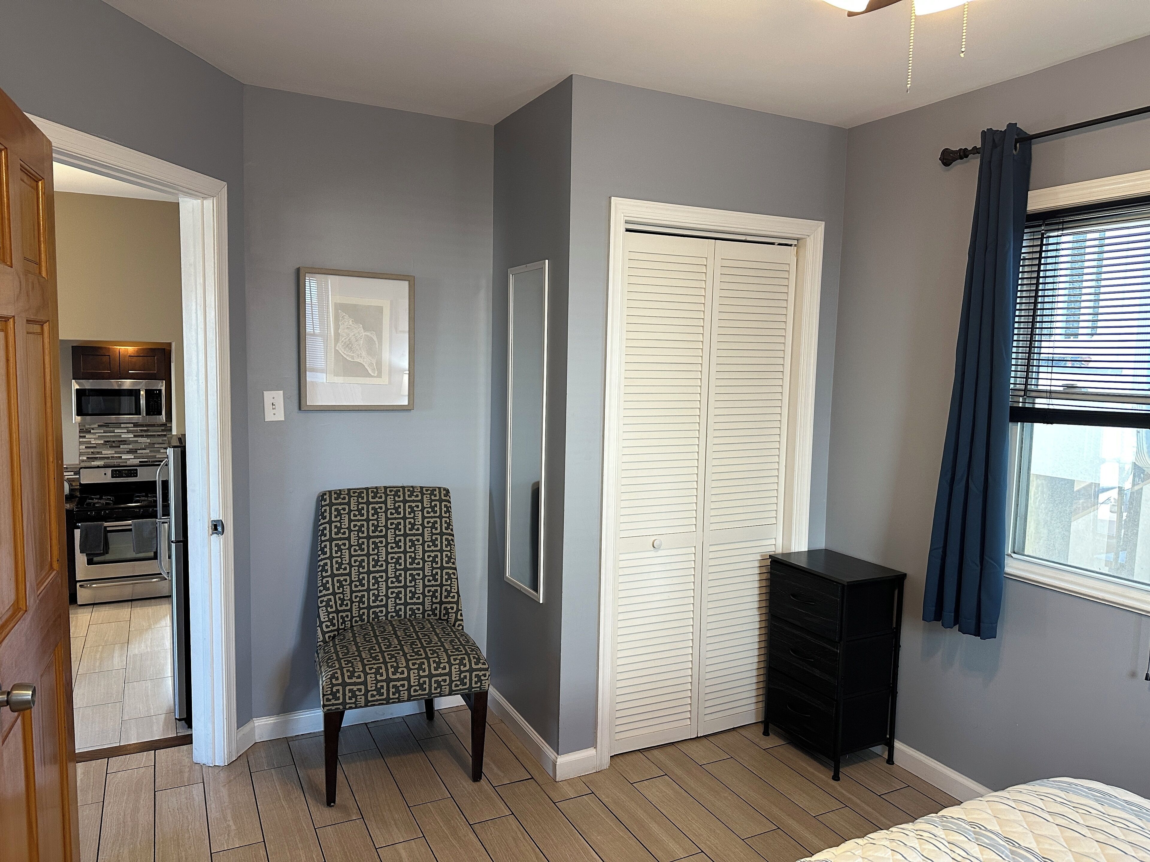 A bedroom with a bed, a wardrobe, a chair, a dresser, and a view of the outside through a window.