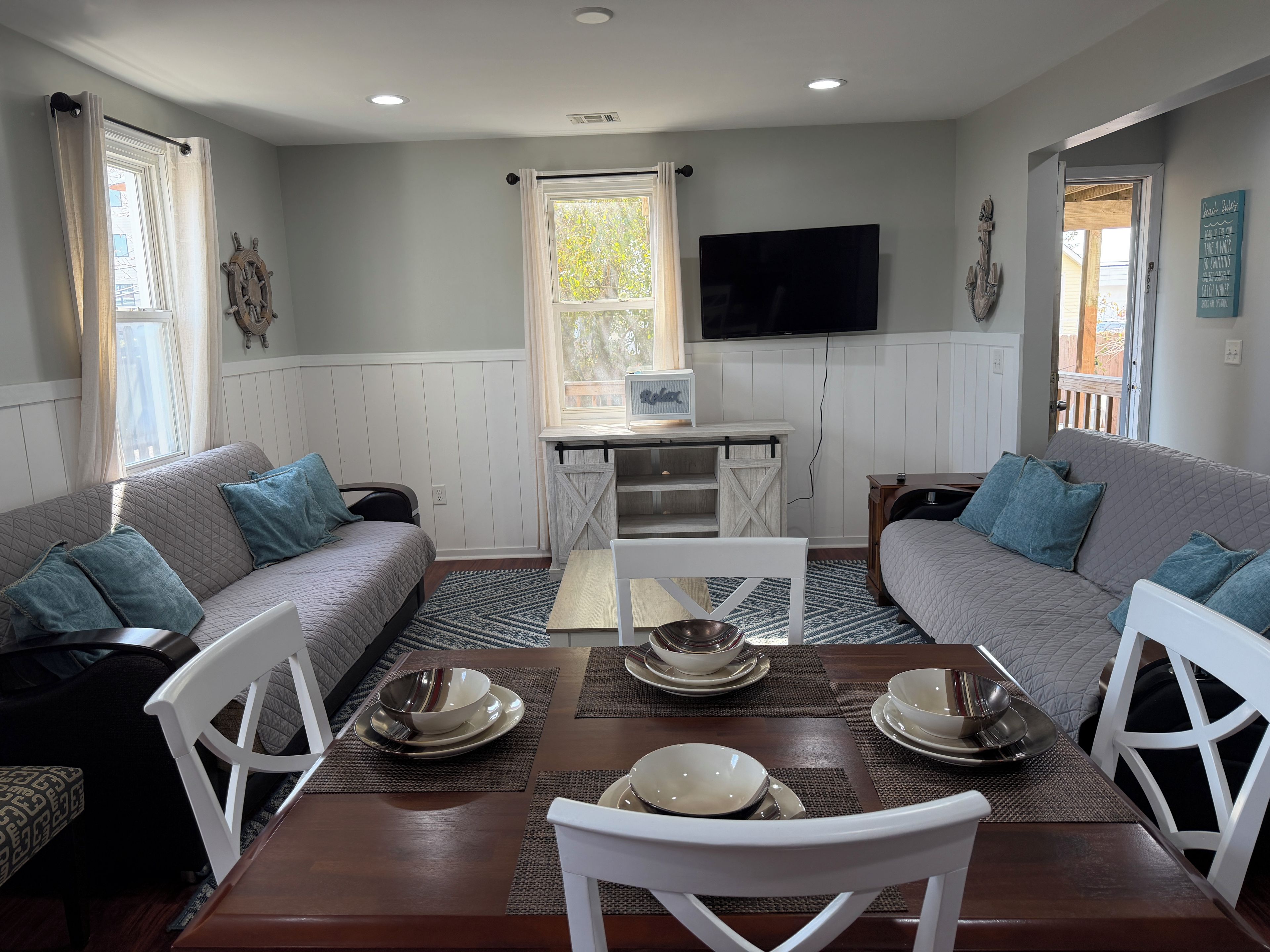 A dining area with a table set for four, surrounded by two sofas, a television mounted on the wall, and a window with curtains.
