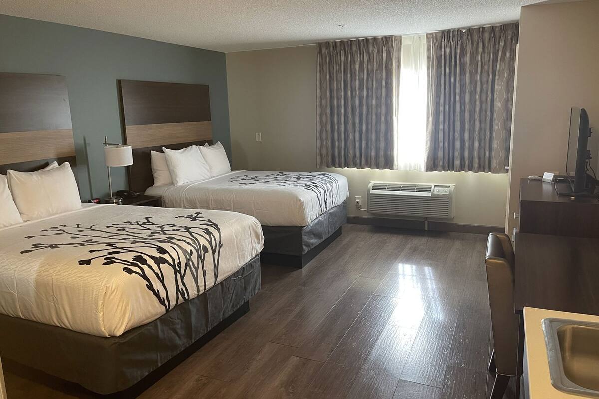 A hotel room with two beds, a desk, a chair, a desk lamp, an air conditioning unit, and a window with curtains.